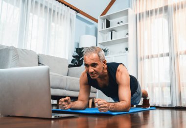 Sportif ve sportif kıdemli bir adam, emekli olduktan sonra sağlıklı yaşam biçimi konsepti olarak ev egzersizinde online fitness videosu izlerken spor paspası takıyor. Etki