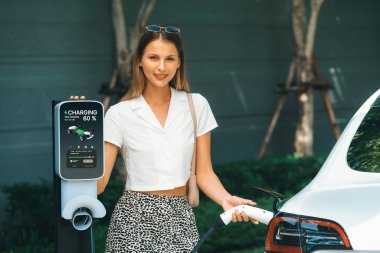 Genç bir kadın yazın yeşil sürdürülebilir şehir bahçesinde EV elektrikli arabasıyla seyahat ediyor. Kentsel sürdürülebilirlik yaşam tarzı yeşil temiz yeniden şarj edilebilir enerji elektrikli BEV araç iç organları