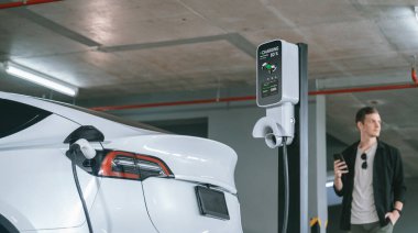 Genç adam EV elektrikli arabasıyla alışveriş merkezinin otoparkına gidiyor şehir merkezindeki şarj oluyor yeşil, temiz, yeniden şarj edilebilir elektrikli araç iç organları ile kentsel sürdürülebilirlik yaşam tarzını gösteriyor
