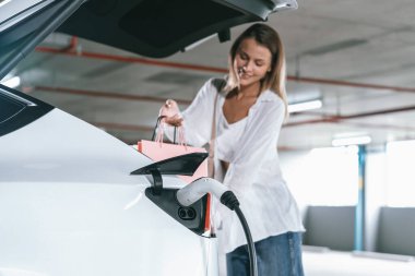 Genç bir kadın EV elektrikli arabasıyla alışveriş merkezinin otoparkına gidiyor şehir merkezinde elektrik iç organlarının yeşil temiz şarj edilebilir enerji ile sürdürülebilirlik yaşam tarzını gösteriyor.