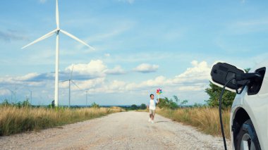 Rüzgar türbini çiftliğinde EV arabasının yanında yel değirmeni oyuncağı oynayan ilerici genç çocuk. Kırsal kesimdeki rüzgar türbini jeneratöründen elektrik jeneratörü..