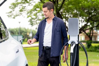Açık yeşil şehir parkı manzaralı EV şarj istasyonundan EV elektrikli araçları şarj eden adam. Çevre dostu şehir ulaşımı ve çevre dostu EV araba seyahati. Yükselt