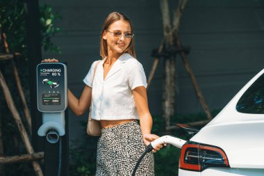 Genç bir kadın yazın yeşil sürdürülebilir şehir bahçesinde EV elektrikli arabasıyla seyahat ediyor. Kentsel sürdürülebilirlik yaşam tarzı yeşil temiz yeniden şarj edilebilir enerji elektrikli BEV araç iç organları