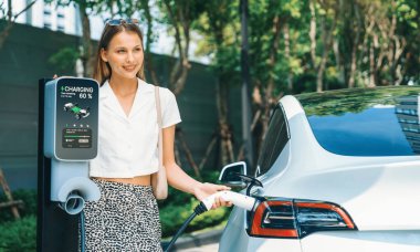 Genç bir kadın yazın yeşil sürdürülebilir şehir bahçesinde EV elektrikli arabasıyla seyahat ediyor. Kentsel sürdürülebilirlik yaşam tarzı yeşil temiz yeniden şarj edilebilir enerji elektrikli BEV araç iç organları