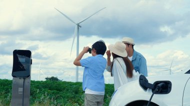 İlerici mutlu aile kavramı elektrikli araçla rüzgar çiftliğinde eğleniyor. Rüzgar türbini jeneratöründen gelen temiz yenilenebilir enerji ile şarj edilen elektrikli araç..