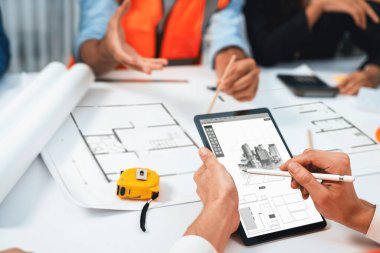 Mimari proje üzerinde birlikte çalışan çeşitli inşaat mühendisi ve müvekkil grupları, toplantı masasında tablet kullanarak inşaat planını gözden geçirip bina planı hazırlıyorlar. Tedbirli