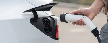 Genç adam şehrin ticari otoparkındaki şarj istasyonundan elektrikli araba aküsünü şarj ediyor. Sürdürülebilir çevre dostu şehir yolculuğu için şarj edilebilir EV araba. Panorama Çıkarı