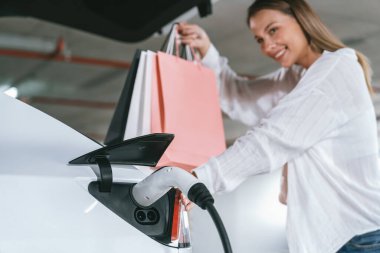 Genç bir kadın EV elektrikli arabasıyla alışveriş merkezinin otoparkına gidiyor şehir merkezinde elektrik iç organlarının yeşil temiz şarj edilebilir enerji ile sürdürülebilirlik yaşam tarzını gösteriyor.