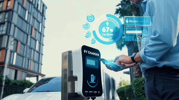 Businessman checking battery status hologram from EV smartphone application while his EV car recharging from charging station in residential area. Futuristic clean sustainable EV tech. Peruse