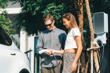 Genç çift yazın yeşil sürdürülebilir şehir bahçesinde EV elektrikli araba ile seyahat ederken, yeşil temiz yeniden şarj edilebilir elektrikli araç iç organları ile kentsel sürdürülebilirlik yaşam tarzını gösteriyor.