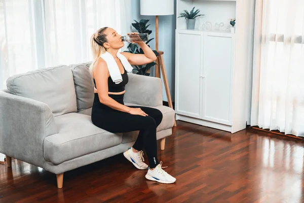 Athletic and sporty senior woman taking a break and sitting on sofa while drinking water after home exercise as concept of healthy fit body lifestyle after retirement. Clout
