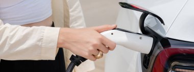 Genç bir kadın şarj aletini şarj istasyonundan elektrikli araba aküsünü şarj etmek için kullanmış. Sürdürülebilir çevre dostu seyahat kavramı için alternatif enerji ve şarj edilebilir EV araba. Panorama Uygulaması