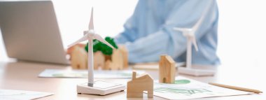 Temiz enerji kullanılarak temsil edilen yel değirmeni modeli ve temsil edilen ahşap blok dizüstü bilgisayarla çalışan iş adamının önünde masaya saçıldı. Geçmişi bulanıklaştırıyor. Tanımlama.