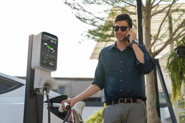 年轻人打电话时在绿城公园停车场给电动车充电 可持续城市生活方式的环保电动车与电池充电站 — 图库照片