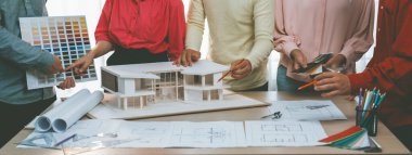 Profesyonel mimar takımının ev rengi seçimi hakkında beyin fırtınası yaparken, mimar da plan ve ev modelinin olduğu masadaki taslak belgeye yazı yazıyor. Değiştirilmiş.