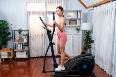 Enerjik ve güçlü atletik Asyalı kadın evde eliptik koşu makinesiyle koşuyor. Formda fiziğin takibi ve ev çalışması ve eğitimle sağlıklı yaşam tarzına bağlılık. Kuvvetli
