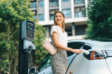 Genç bir kadın yazın yeşil sürdürülebilir şehir bahçesinde EV elektrikli arabasıyla seyahat ediyor. Kentsel sürdürülebilirlik yaşam tarzı yeşil temiz yeniden şarj edilebilir enerji elektrikli BEV araç iç organları