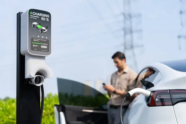 Man pay for electricity with smartphone while recharge EV car battery at charging station connected to power grid tower electrical as electrical industry for eco friendly car utilization.Expedient