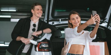 Genç çift EV elektrikli arabasıyla alışveriş merkezinin otoparkına gidiyor şehir merkezindeki şarj oluyor yeşil temiz şarj edilebilir elektrikli araç iç organları ile kentsel sürdürülebilirlik yaşam tarzını gösteriyor