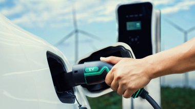 EV şarj aletini elle yerleştirin ve rüzgar türbini jeneratörü arka planına sahip şarj istasyonundan elektrikli arabayı şarj edin. Zeki çevre dostu EV araba. Alternatif enerjinin teknolojik gelişimi. İnceleyin