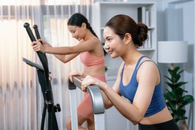 Enerjik ve güçlü atletik Asyalı kadın evde spor arkadaşı ya da antrenörü ile birlikte eliptik koşu makinesiyle koşuyor. Formda fiziğin takibi ve sağlıklı yaşam tarzına bağlılık. Kuvvetli