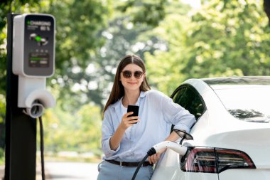 Ulusal Park veya Yaz Ormanı 'ndaki tatil gezisi sırasında EV şarj istasyonundan elektrik aküsü almak için akıllı telefon internet bankacılık uygulaması kullanan genç bir kadın. Yükselt