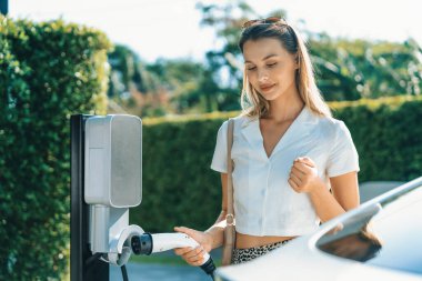 Genç bir kadın yazın yeşil sürdürülebilir şehir bahçesinde EV elektrikli arabasıyla seyahat ediyor. Kentsel sürdürülebilirlik yaşam tarzı yeşil temiz yeniden şarj edilebilir enerji elektrikli BEV araç iç organları