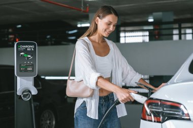 Genç bir kadın EV elektrikli arabasıyla alışveriş merkezinin otoparkına gidiyor şehir merkezinde elektrik iç organlarının yeşil temiz şarj edilebilir enerji ile sürdürülebilirlik yaşam tarzını gösteriyor.