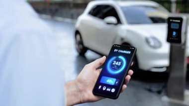 Akıllı telefon ekran bataryasını elinde tutan iş adamı akıllı EV mobil uygulama ile otomobil parkındaki şarj istasyonundan elektrik şarj eden çevre dostu EV otomobil. İnceleyin