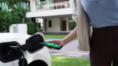 Modern yaşam tarzı bir kadın EV şarj cihazını ev şarj istasyonuna yerleştirir ve akıllı telefondan EV batarya durumunu etkinleştirir. Gelecekçi enerji sürdürülebilirliği ve elektrikli araba. İnceleyin