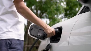 Adam EV şarj aleti takıyor ve yeşil park ve yeşillik arka planında akıllı şarj istasyonu olan elektrikli arabayı şarj ediyor. Çevre koruma için temiz enerjiden güç alan çevre dostu EV araba. İnceleyin
