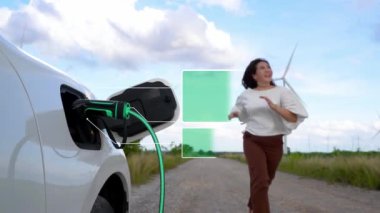 Çevre dostu EV otomobili ekran durumu hologramını şarj ediyor, çevreye duyarlı rüzgar türbini jeneratörüyle şarj ediyor ve arka planda mutlu bir aile uçurtması oynuyor. İnceleyin