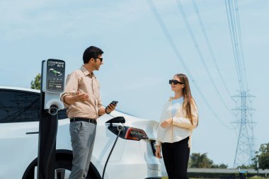 Couple pay for electricity with smartphone while recharge EV car battery at charging station connected to power grid tower electrical as electrical industry for eco friendly car utilization.Expedient clipart