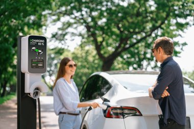Doğal orman ya da milli parktaki araba yolculuğu sırasında elektrikli araba için şarj olan güneş gözlüğü takan sevimli genç bir çift. Tatil ve tatil boyunca çevre dostu seyahat. Yükselt