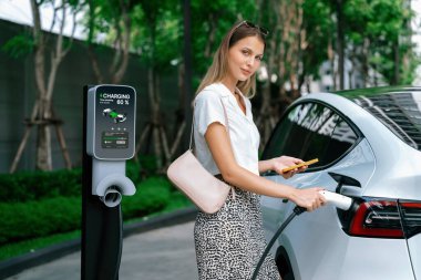 Genç bir kadın yazın yeşil sürdürülebilir şehir bahçesinde EV elektrikli arabasıyla seyahat ediyor. Kentsel sürdürülebilirlik yaşam tarzı yeşil temiz yeniden şarj edilebilir enerji elektrikli BEV araç iç organları