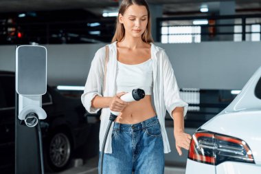 Genç bir kadın EV elektrikli arabasıyla alışveriş merkezinin otoparkına gidiyor şehir merkezinde elektrik iç organlarının yeşil temiz şarj edilebilir enerji ile sürdürülebilirlik yaşam tarzını gösteriyor.
