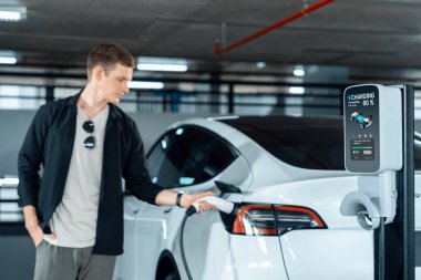 Genç adam EV elektrikli arabasıyla alışveriş merkezinin otoparkına gidiyor şehir merkezindeki şarj oluyor yeşil, temiz, yeniden şarj edilebilir elektrikli araç iç organları ile kentsel sürdürülebilirlik yaşam tarzını gösteriyor