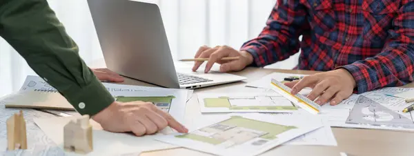 Professional architect design a blueprint by using laptop during project manager shows mistake point on table with architectural document and wooden block scatter around.. Closeup. Delineation.