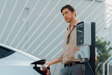 Genç adam, şehrin ticari otoparkındaki şarj istasyonundan elektrikli araba aküsünü şarj etmek için EV şarj aleti koymuş. Sürdürülebilir çevre dostu şehir yolculuğu için şarj edilebilir EV araba. Yararlı