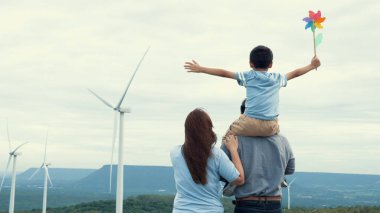 İlerici mutlu aile kavramı rüzgâr türbini çiftliğinde eğleniyor. Rüzgar türbini jeneratörüyle gelen elektrik jeneratörü kırsal bölgede tepe ve ufukta dağ var..