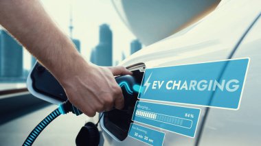 EV şarj aletini elle yerleştirin ve şarj istasyonundan elektrikli arabayı şarj edin. Şehrin bulanık arka planında gelecekteki batarya durum hologramını görüntüleyin. Akıllı sürdürülebilir temiz enerji. İnceleyin