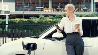 Şehir merkezinde dolaşmadan önce elektrikli araba ve şarj istasyonuna yaslanan ilerici iş kadını. Çevre dostu şarj edilebilir EV araba sürdürülebilir ve temiz enerjiyle çalışıyor.
