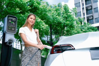 Genç bir kadın yazın yeşil sürdürülebilir şehir bahçesinde EV elektrikli arabasıyla seyahat ediyor. Kentsel sürdürülebilirlik yaşam tarzı yeşil temiz yeniden şarj edilebilir enerji elektrikli BEV araç iç organları