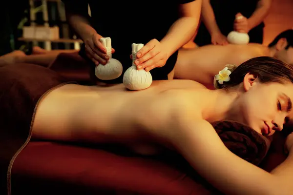 Hot herbal ball spa massage body treatment, masseur gently compresses herb bag on couple customer body. Serenity of aromatherapy recreation in warm lighting of candles at spa salon. Quiescent