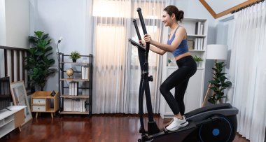 Enerjik ve güçlü atletik Asyalı kadın evde eliptik koşu makinesiyle koşuyor. Formda fiziğin takibi ve ev çalışması ve eğitimle sağlıklı yaşam tarzına bağlılık. Kuvvetli