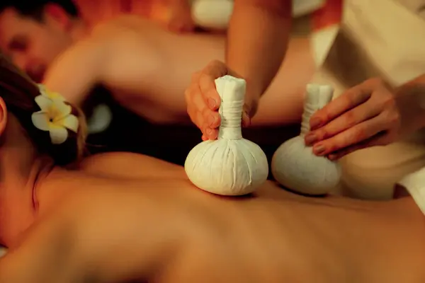Closeup hot herbal ball spa massage body treatment, masseur gently compresses herb bag on couple customer body. Serenity of aromatherapy recreation in warm lighting of candles at spa salon. Quiescent