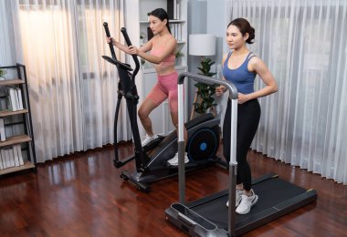 Enerjik ve güçlü atletik Asyalı kadın evde spor arkadaşı ya da antrenörü ile birlikte eliptik koşu makinesiyle koşuyor. Formda fiziğin takibi ve sağlıklı yaşam tarzına bağlılık. Kuvvetli