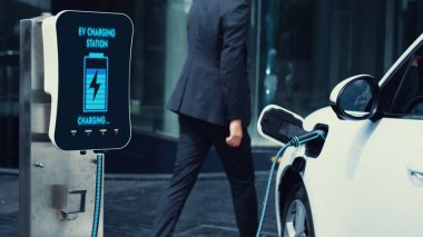 İş adamı elektrikli arabasını şehir merkezindeki şarj istasyonundan ya da otoparktan şarj ediyor. Şehir yaşam tarzında alternatif temiz enerji kullanan çevre dostu şarj edilebilir araba. İnceleyin.