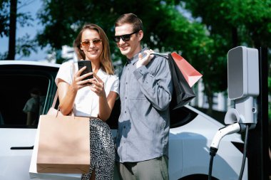 Genç çift yazın yeşil sürdürülebilir şehir bahçesinde EV elektrikli araba ile seyahat ederken, yeşil temiz yeniden şarj edilebilir elektrikli araç iç organları ile kentsel sürdürülebilirlik yaşam tarzını gösteriyor.