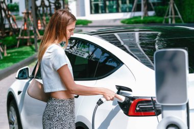 Genç bir kadın yazın yeşil sürdürülebilir şehir bahçesinde EV elektrikli arabasıyla seyahat ediyor. Kentsel sürdürülebilirlik yaşam tarzı yeşil temiz yeniden şarj edilebilir enerji elektrikli BEV araç iç organları